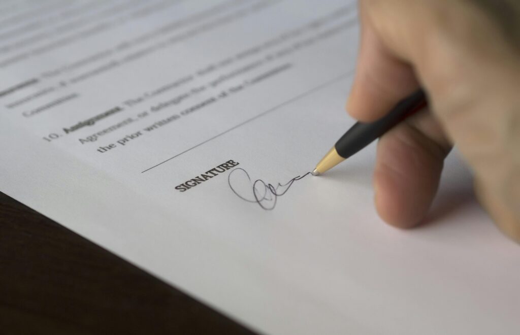 A hand signs a formal contract with a pen on a wooden desk.